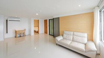 A modern living room with a white sofa, a wooden dining table, and a glass door leading to a bedroom.