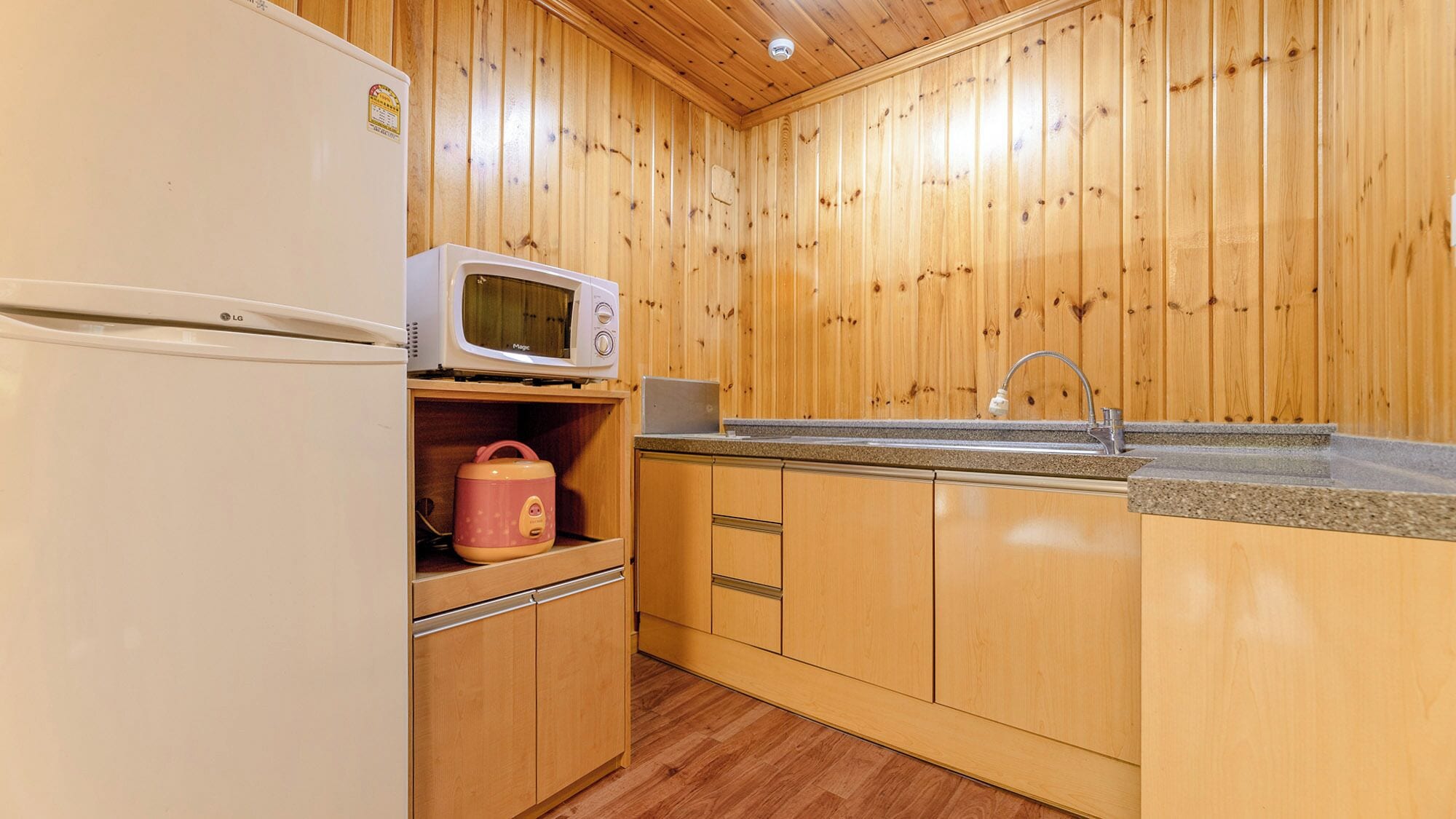 A compact kitchen with wooden walls, a white refrigerator, a microwave, a sink, and a pink kettle.
