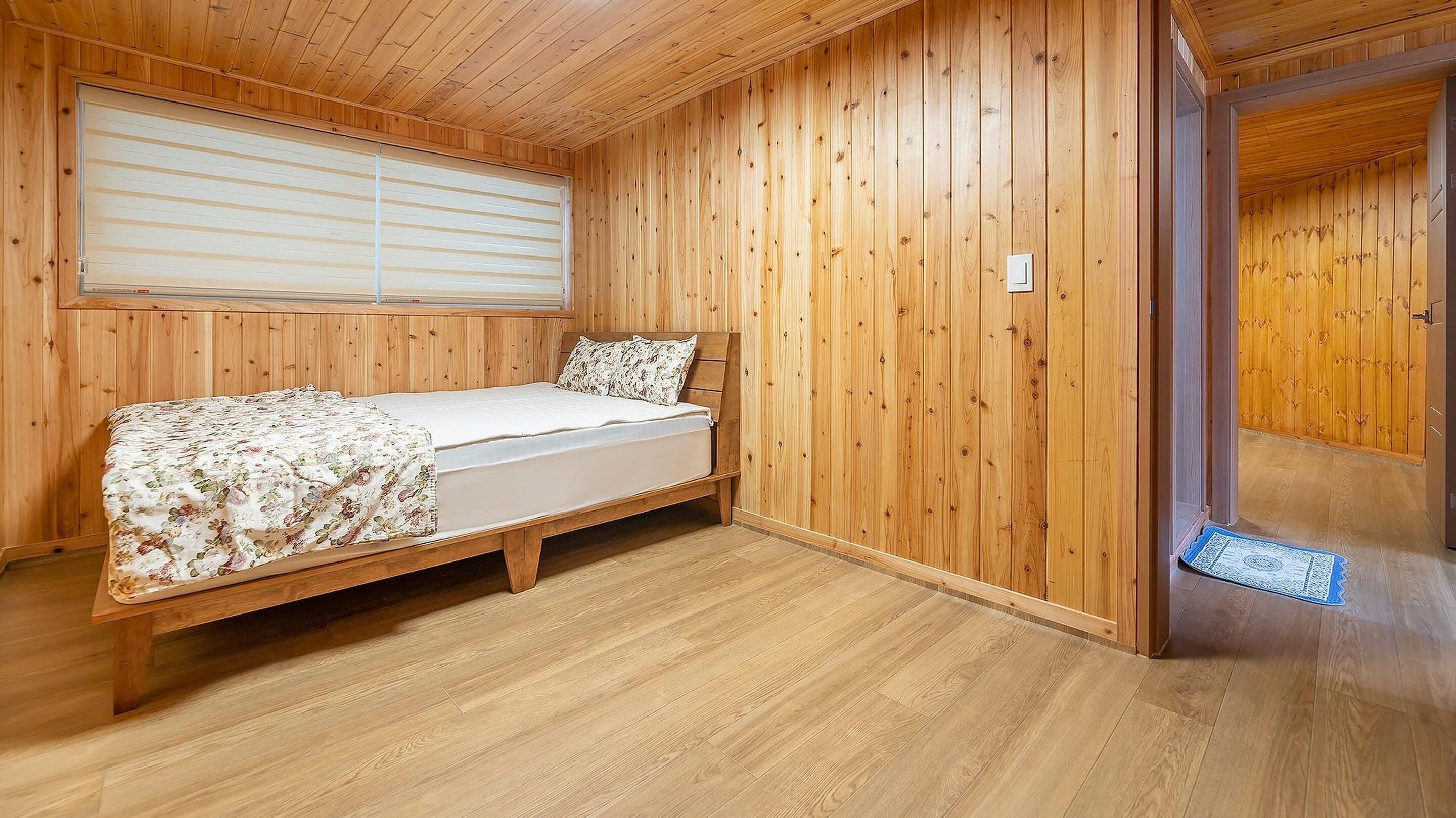 A wooden-paneled room with a bed, a window with blinds, and a door leading to another room.