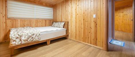 A wooden-paneled room with a bed, a window with blinds, and a door leading to another room.
