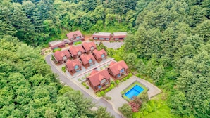 A cluster of red-roofed buildings surrounded by dense green forest, with a swimming pool in the foreground.