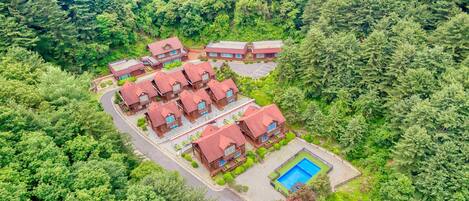 Un conjunto de edificios con techos rojos rodeado de un bosque frondoso, con una piscina en primer plano.