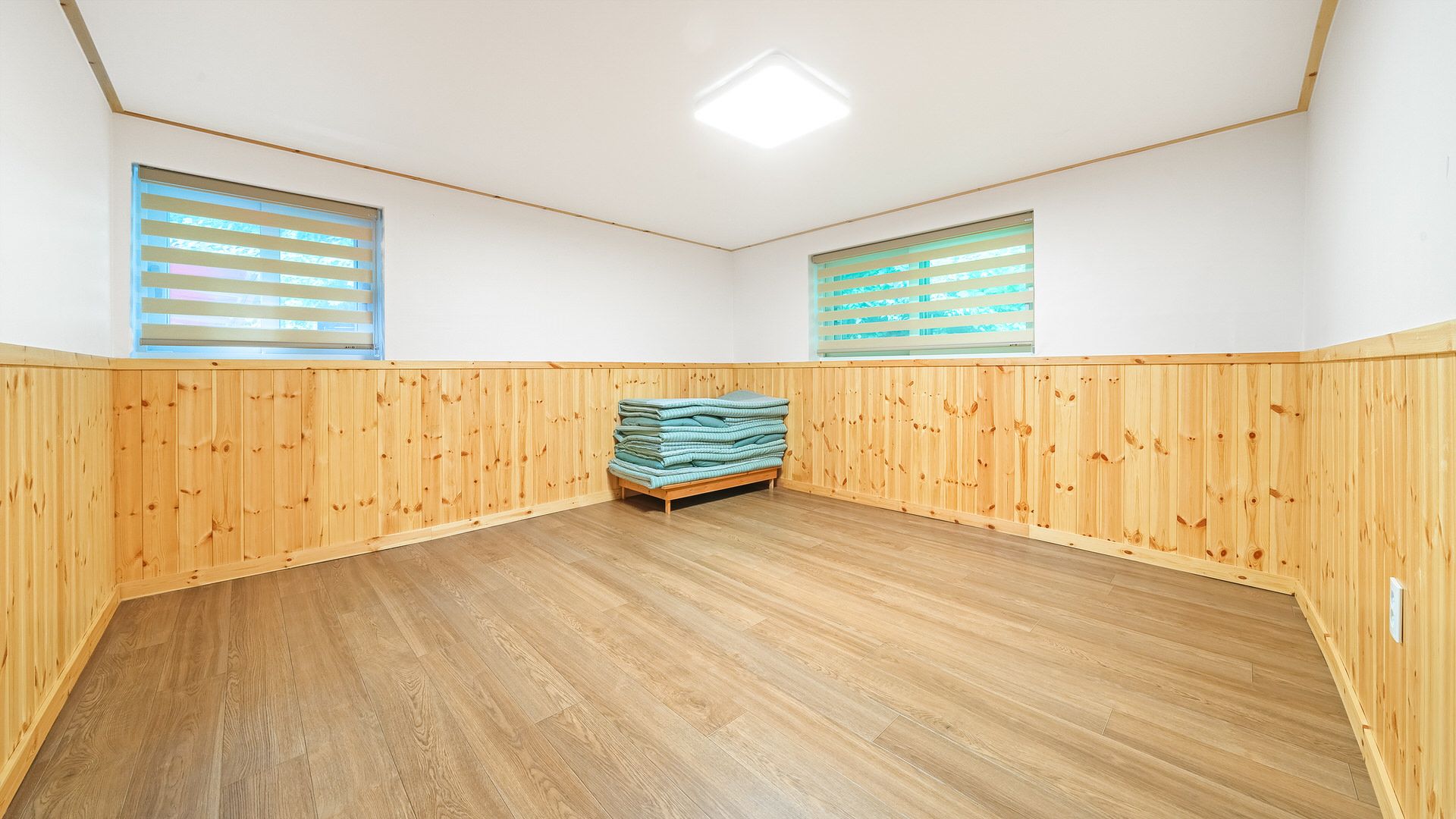 A room with wooden panel walls, a central bed, and two windows with blinds.