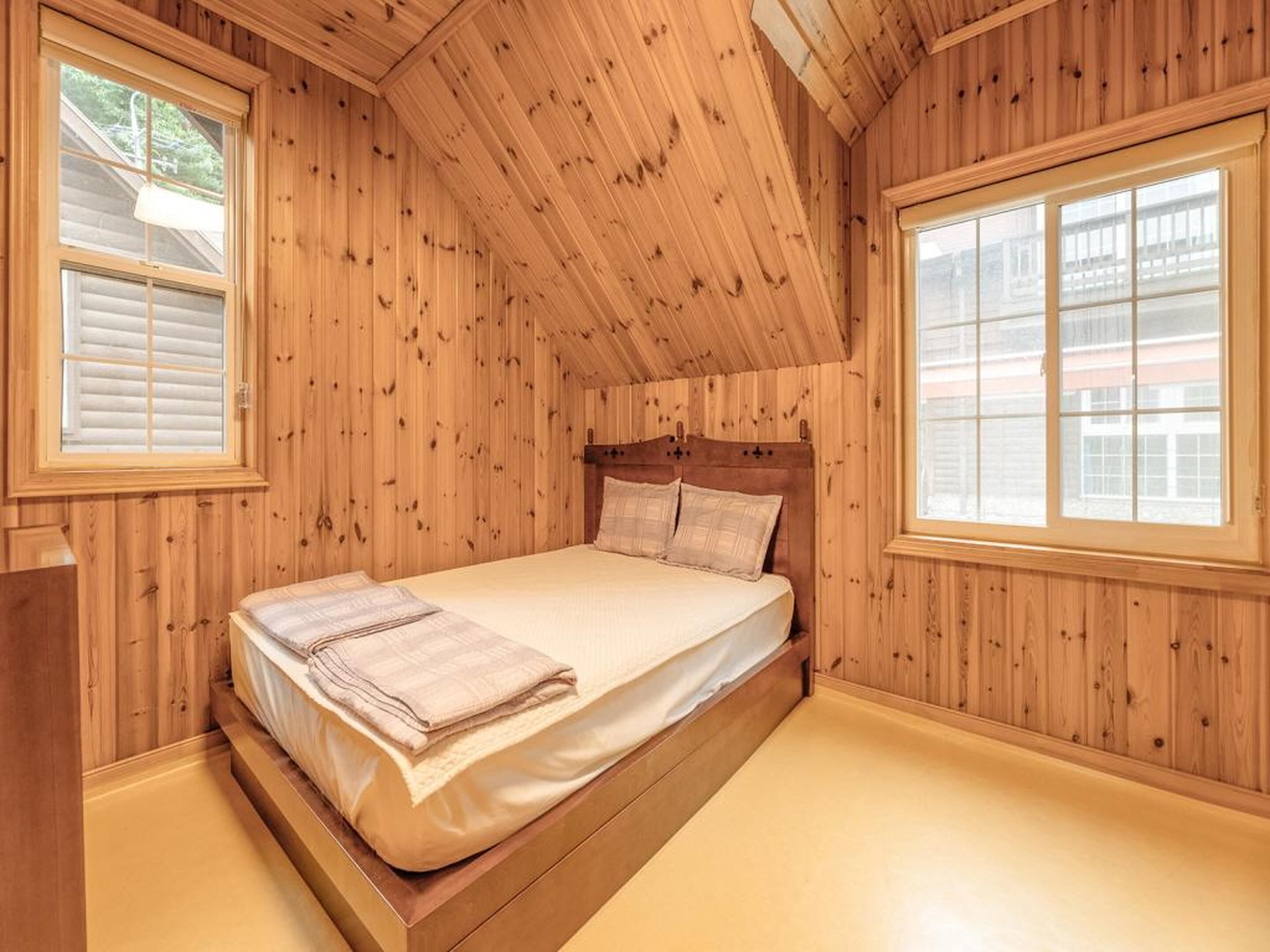 Un dormitorio con paneles de madera, una cama individual, dos ventanas y un techo inclinado.