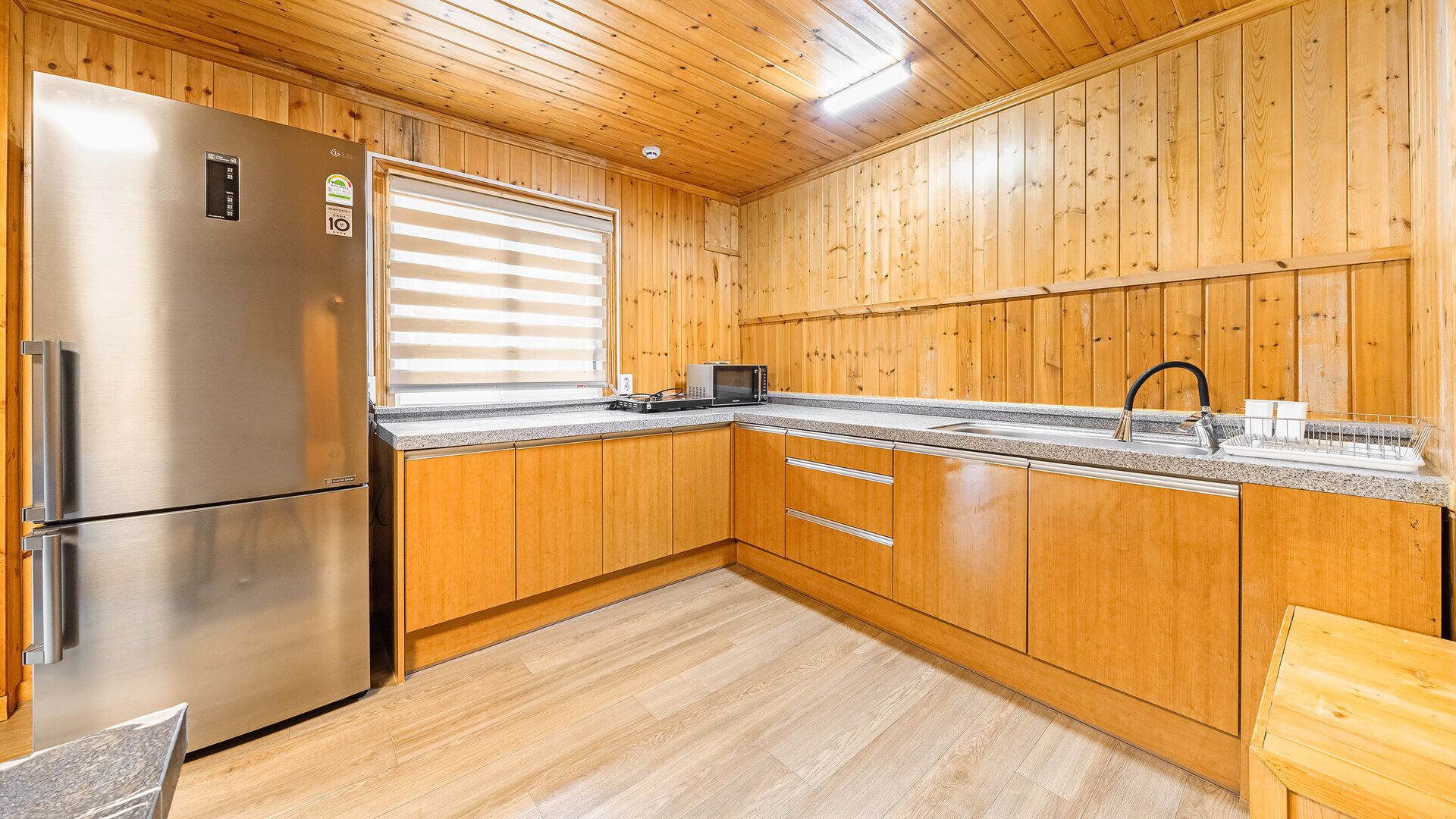 A kitchen with wooden cabinets, a stainless steel refrigerator, a microwave, and a sink with a dish rack.