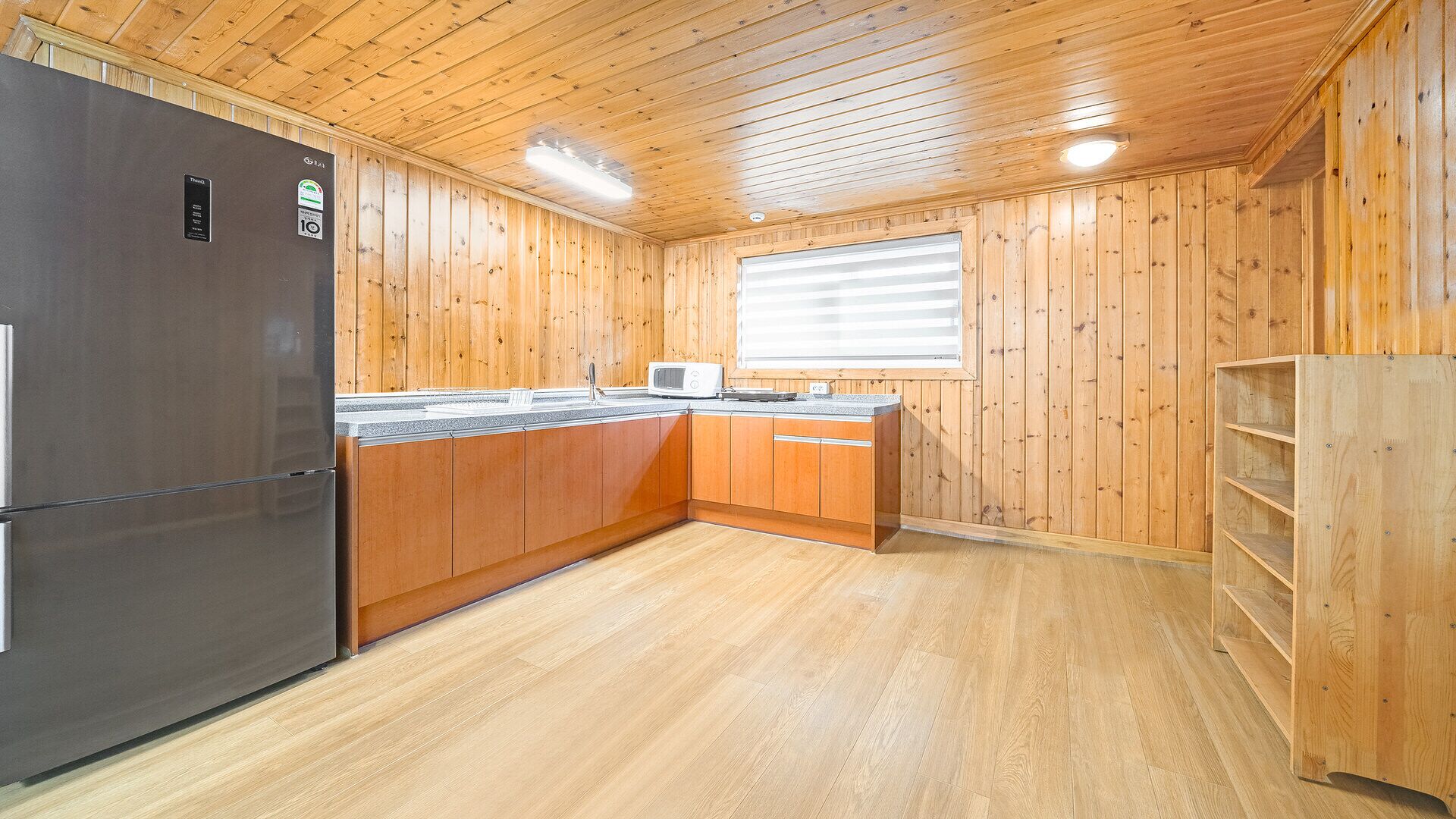 Une cuisine compacte aux murs en bois, équipée d’un réfrigérateur, d’un four à micro-ondes et d’une fenêtre avec des stores.