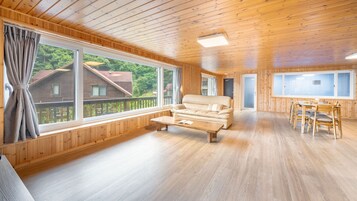 Habitación amplia con paredes y techo de madera, un sofá, una mesa de comedor con sillas, y ventanas grandes que ofrecen vistas a una casa y árboles.