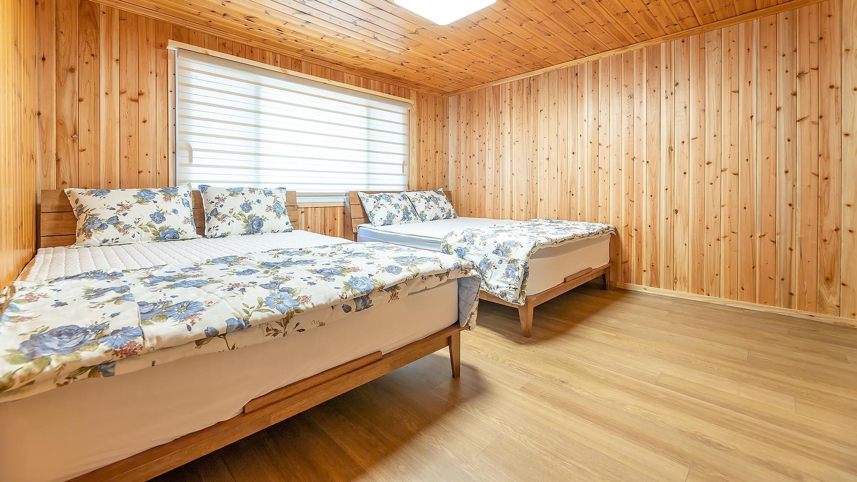Une chambre avec deux lits, des murs en bois, une grande fenêtre avec des stores et un plafond en pente.