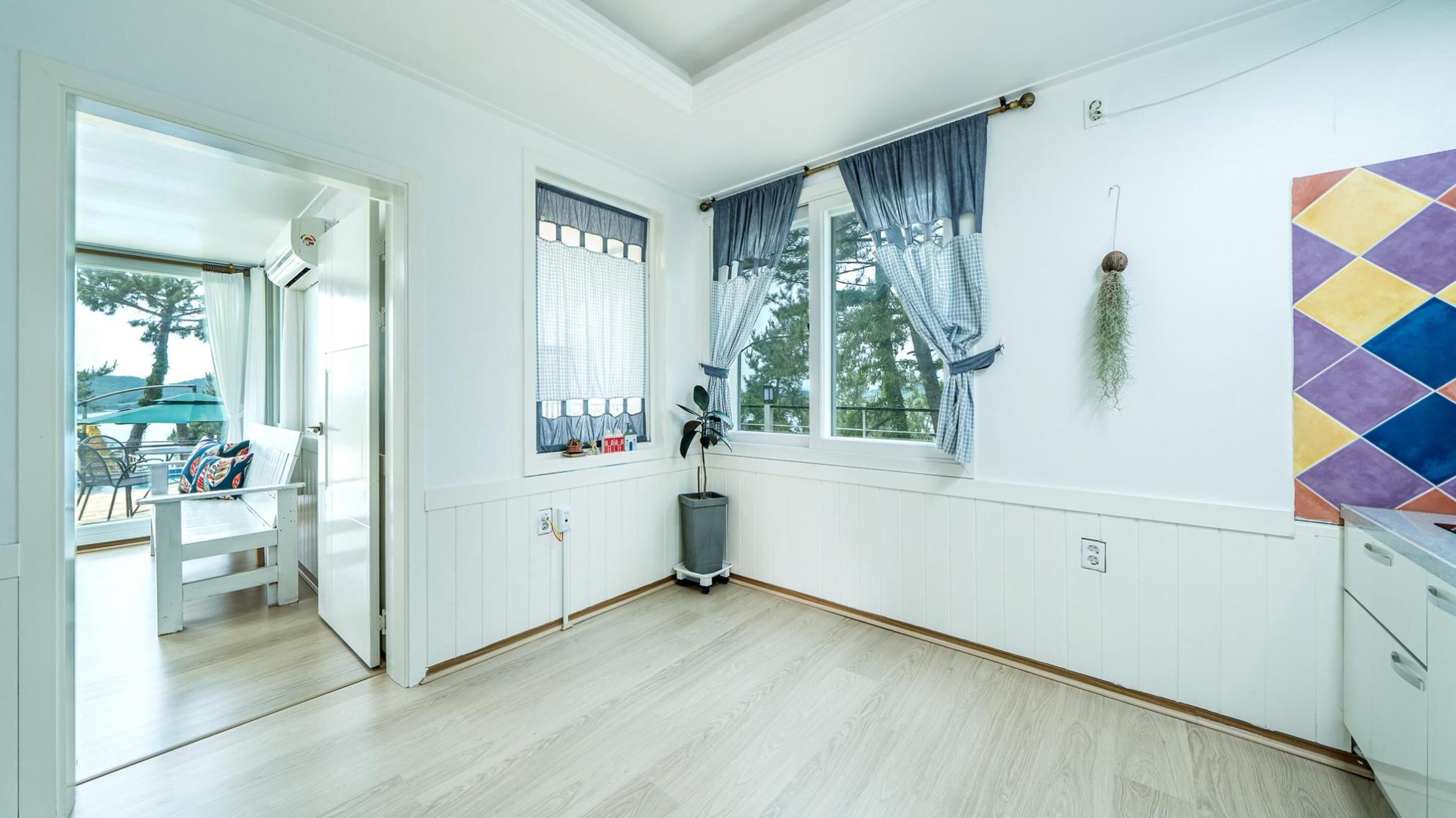 Een kamer met houten vloer, witte wanden, een raam met blauwe gordijnen, een plant in pot en een kleurrijke decoratie aan de muur.