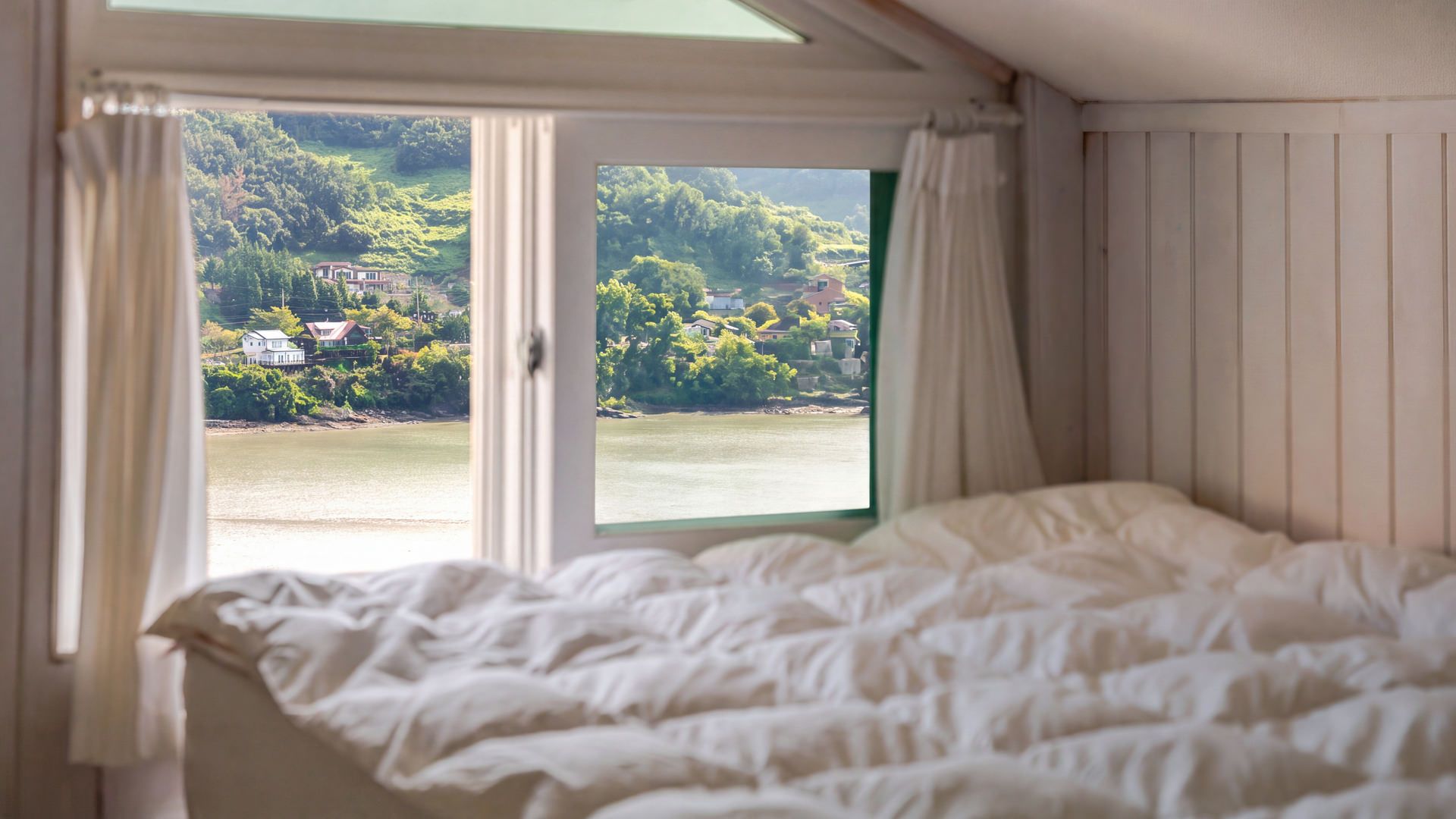 A bedroom with a bed, a window with curtains, and a view of a lake and houses.