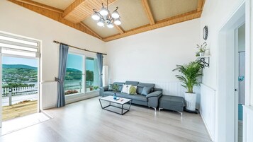 A spacious living room with a large window, a grey sofa, a coffee table, and a ceiling with wooden beams and a chandelier.