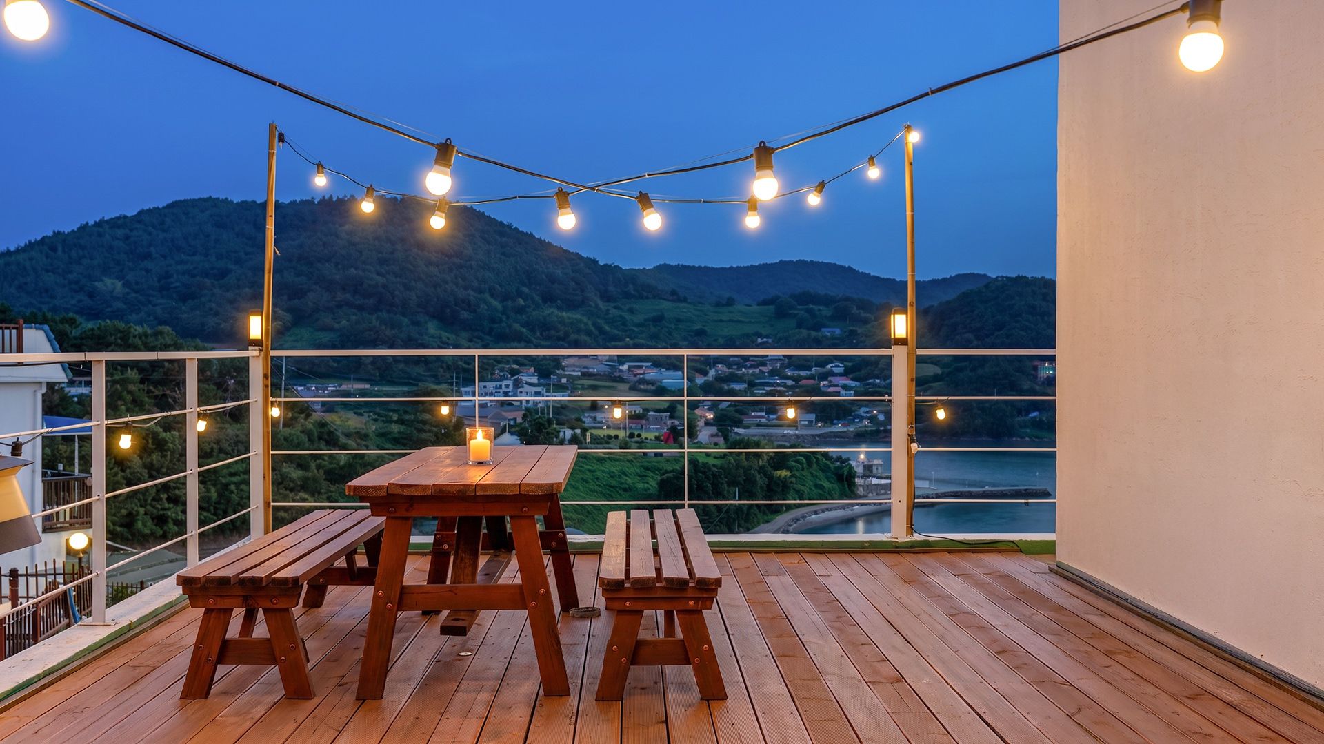 A wooden deck with a table and benches, string lights, and a view of a town and mountains.