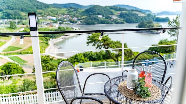 A balcony with a table set for two, overlooking a river and hills. - Yeosu Sky Pension (Yeosu)