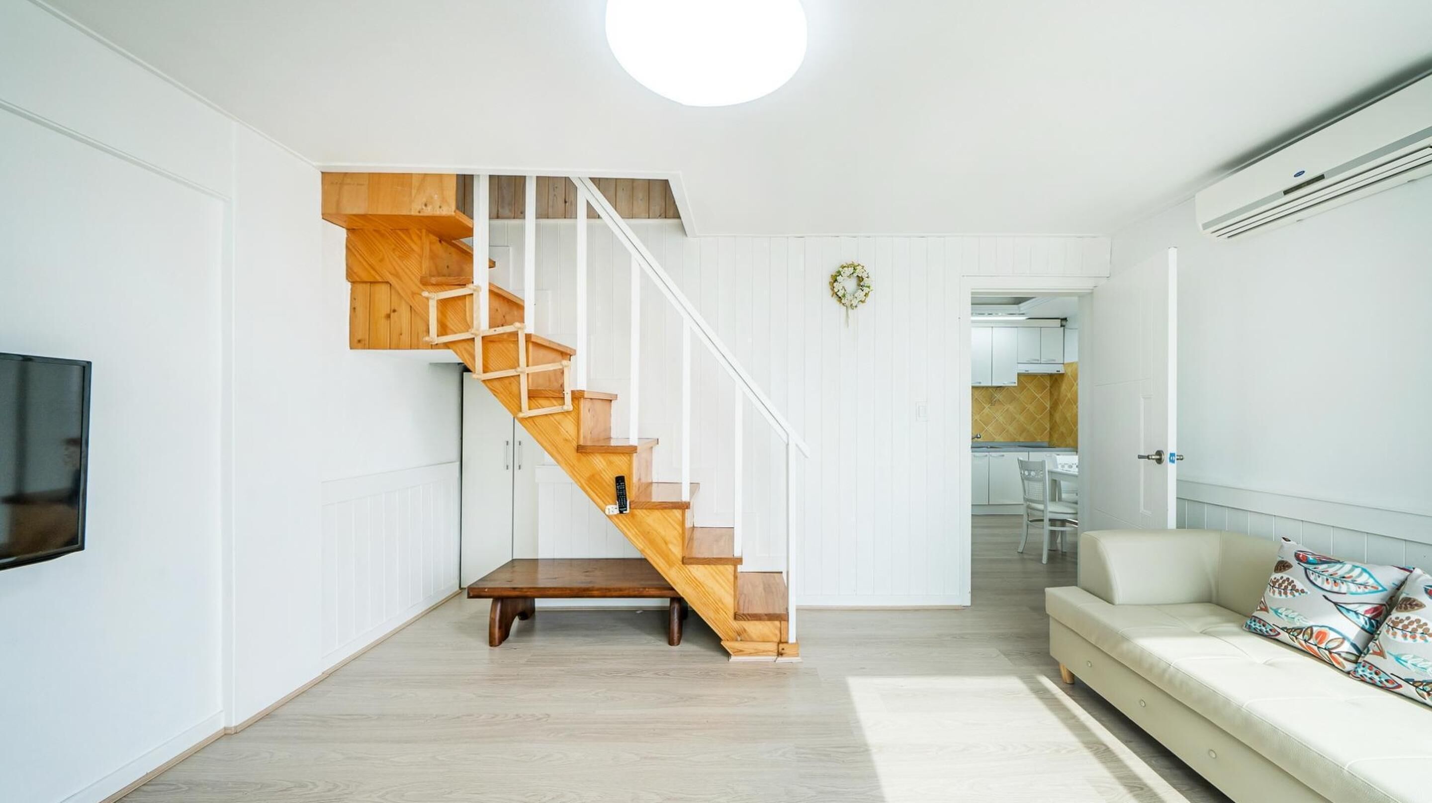 Un salon moderne avec un escalier en bois, un téléviseur à écran plat, un canapé agrémenté de coussins à motifs et une cuisine visible à travers une porte ouverte.