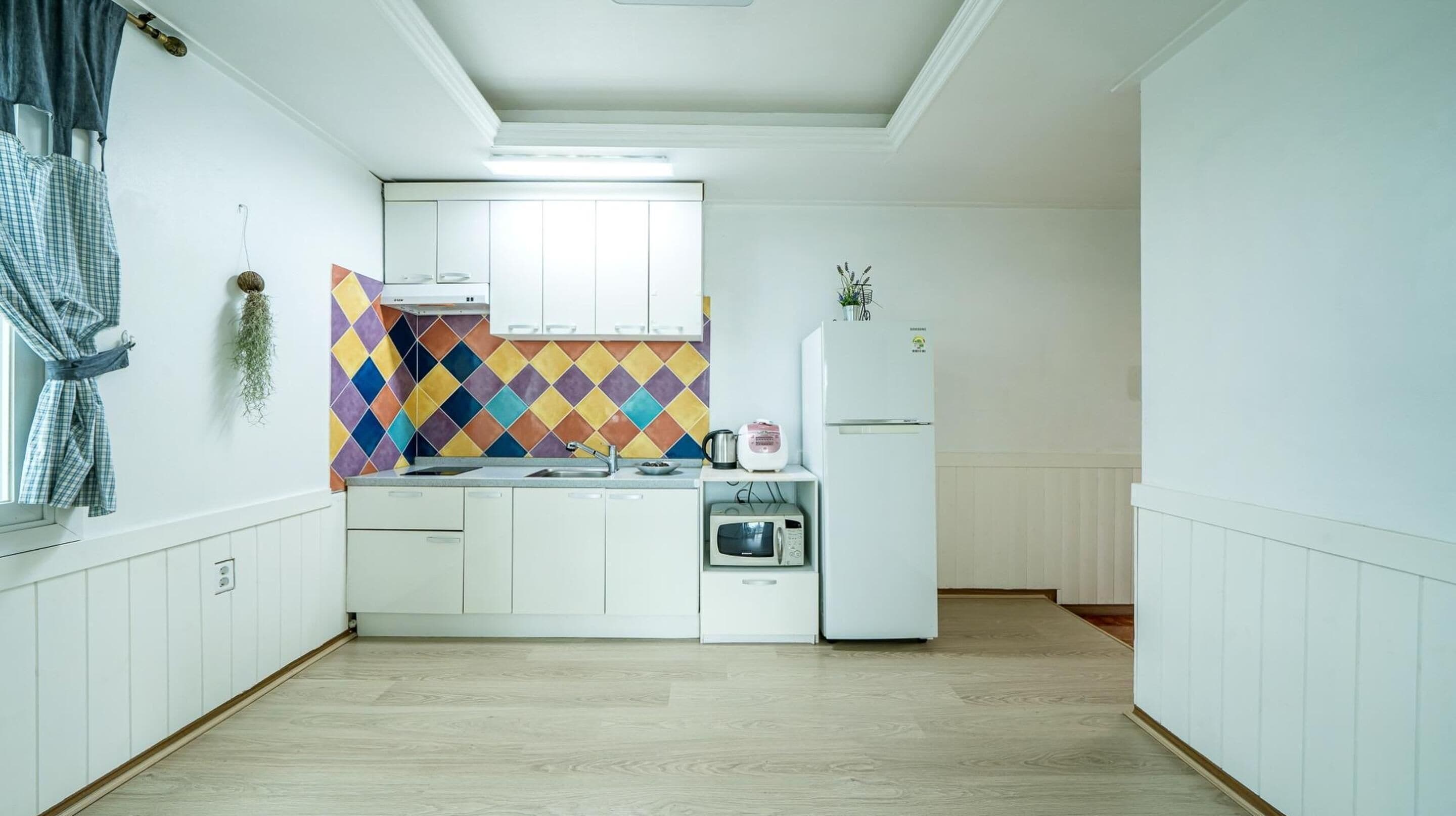 A compact kitchen with white cabinets, a colorful diamond-patterned backsplash, a microwave, a kettle, and a refrigerator.