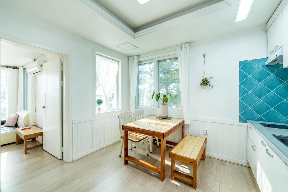 Eine helle Küche mit einem Holztisch und einer Bank, einer blauen Fliesenrückwand und einem Fenster mit Blick auf Bäume.