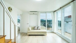 Un salón espacioso con sofá, un balcón con mesa y sillas, y ventanas amplias con vistas al exterior.