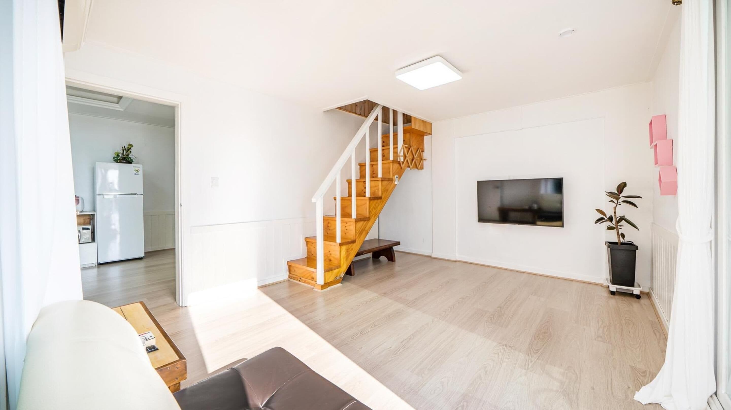 A spacious living area with a wooden staircase, a flat-screen TV mounted on the wall, and a potted plant.