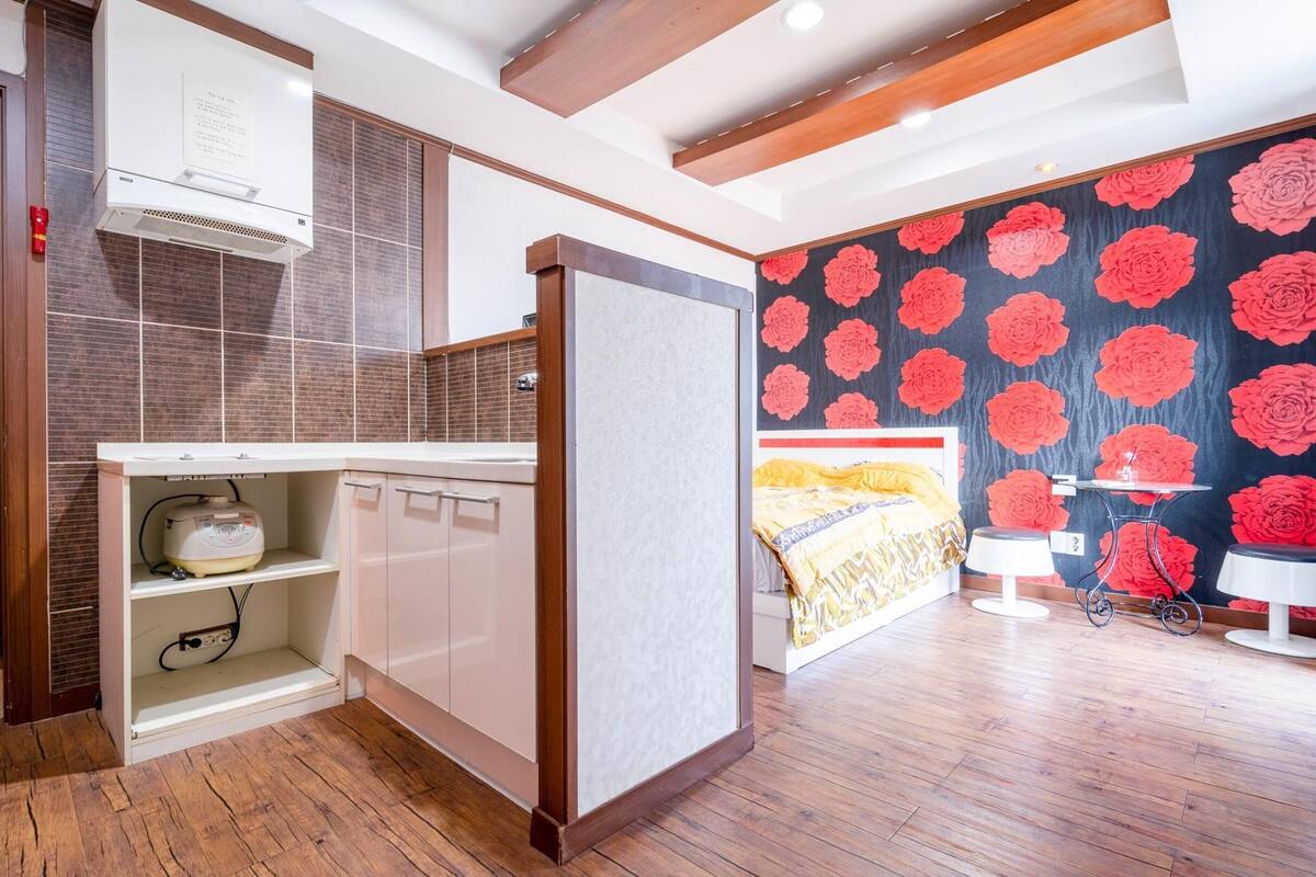 Une chambre d’hôtel avec un plancher en bois, un lit recouvert d’une couvre-lit à motifs jaunes et blancs, une table de chevet avec une lampe, et un mur orné d’un papier peint à motifs floraux.
