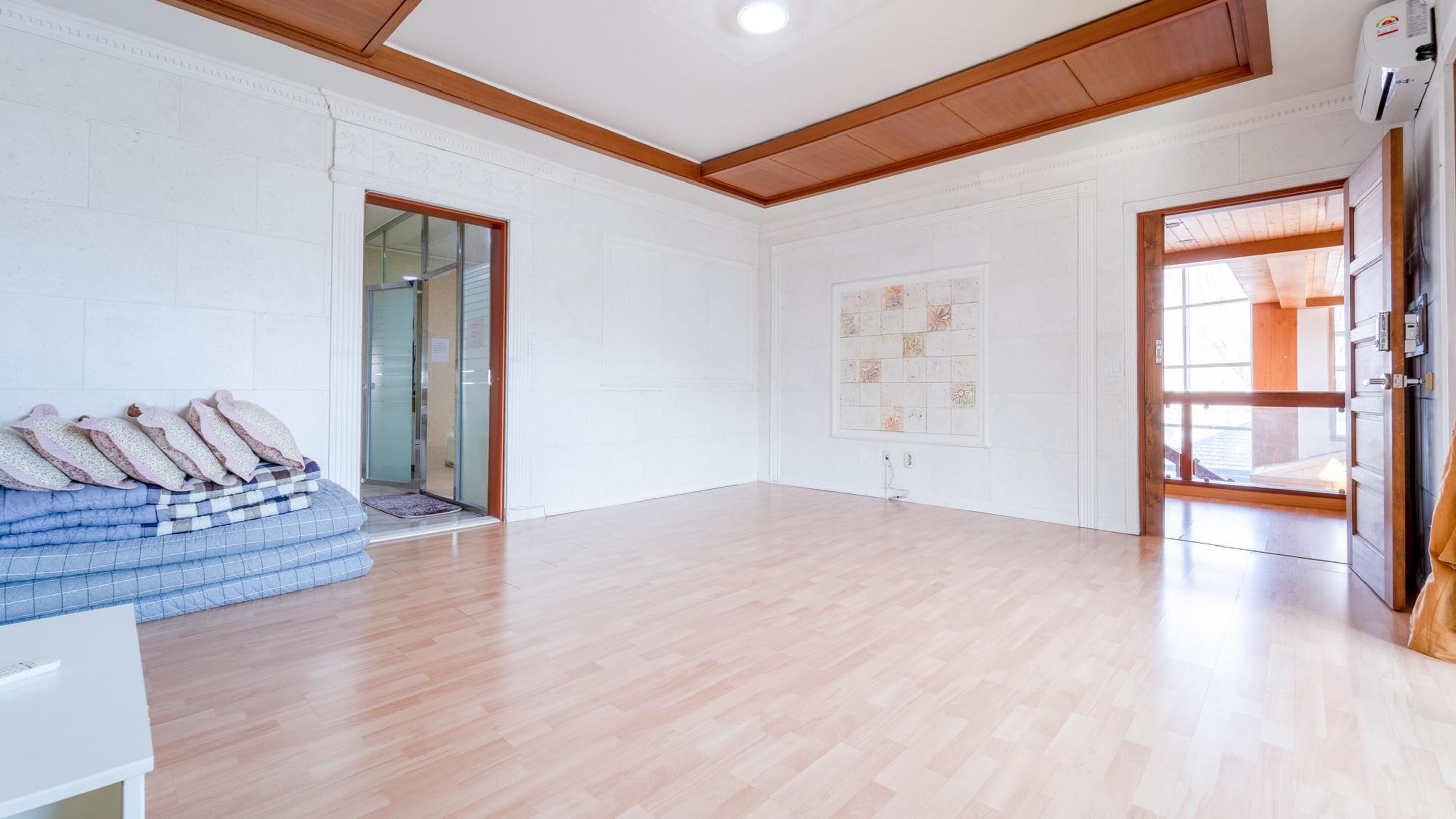 Une pièce spacieuse avec un plancher en bois, un mur blanc orné de moulures décoratives, une grande fenêtre et une porte donnant sur l’extérieur.
