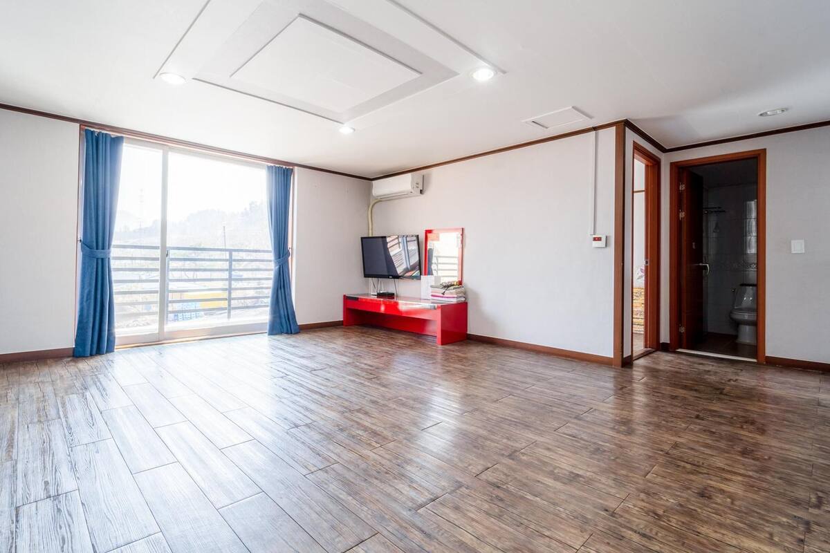 Une pièce spacieuse avec un plancher en bois, une grande fenêtre ornée de rideaux bleus, un téléviseur à écran plat posé sur un meuble rouge, et une porte de salle de bain visible.