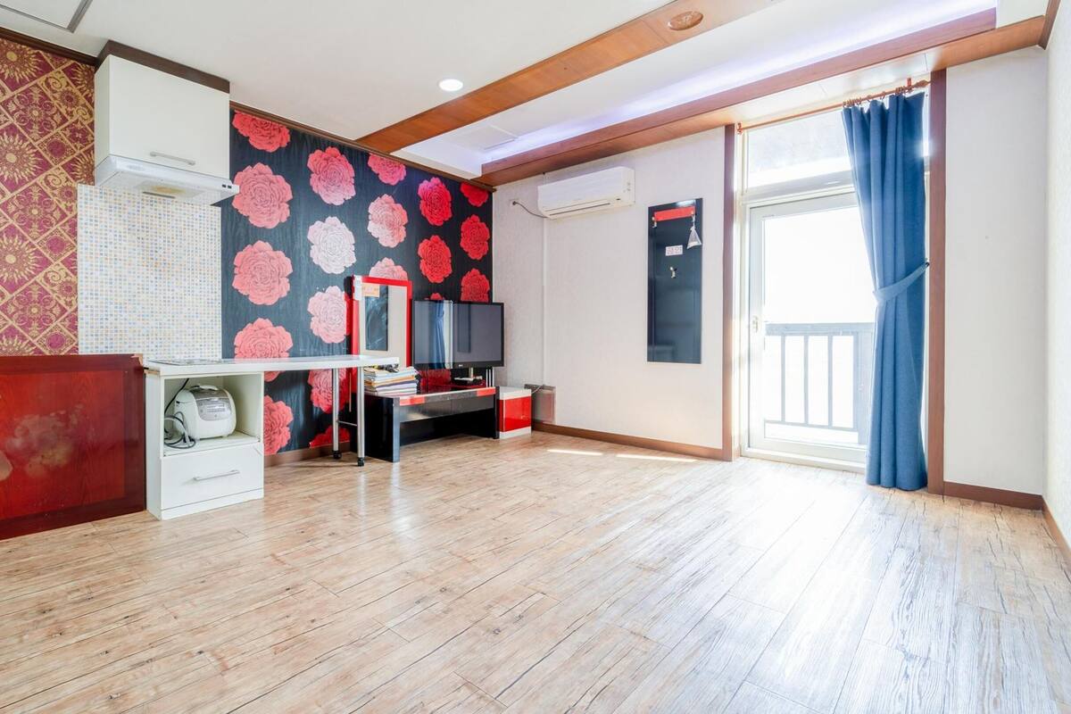 Une pièce avec un plancher en bois, un buffet rouge, un réfrigérateur blanc, une télévision et une grande fenêtre ornée de rideaux bleus.