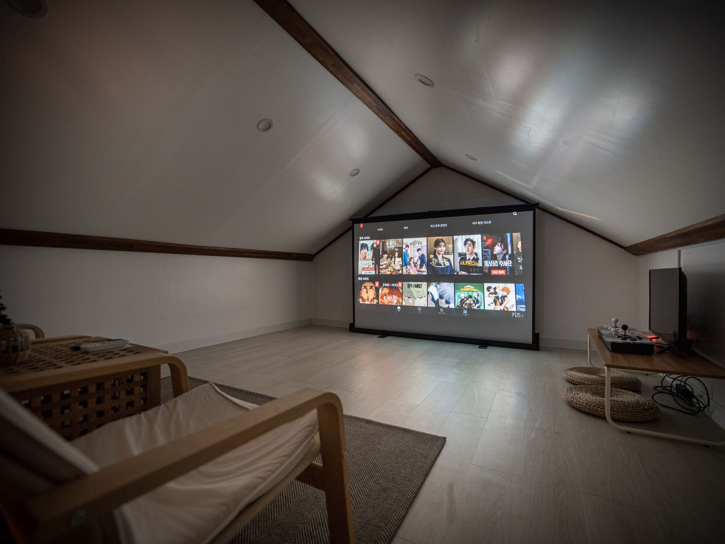 A modern, sparsely furnished room with a sloped ceiling, a large flat-screen TV displaying a streaming service interface, and a small table with a remote control.