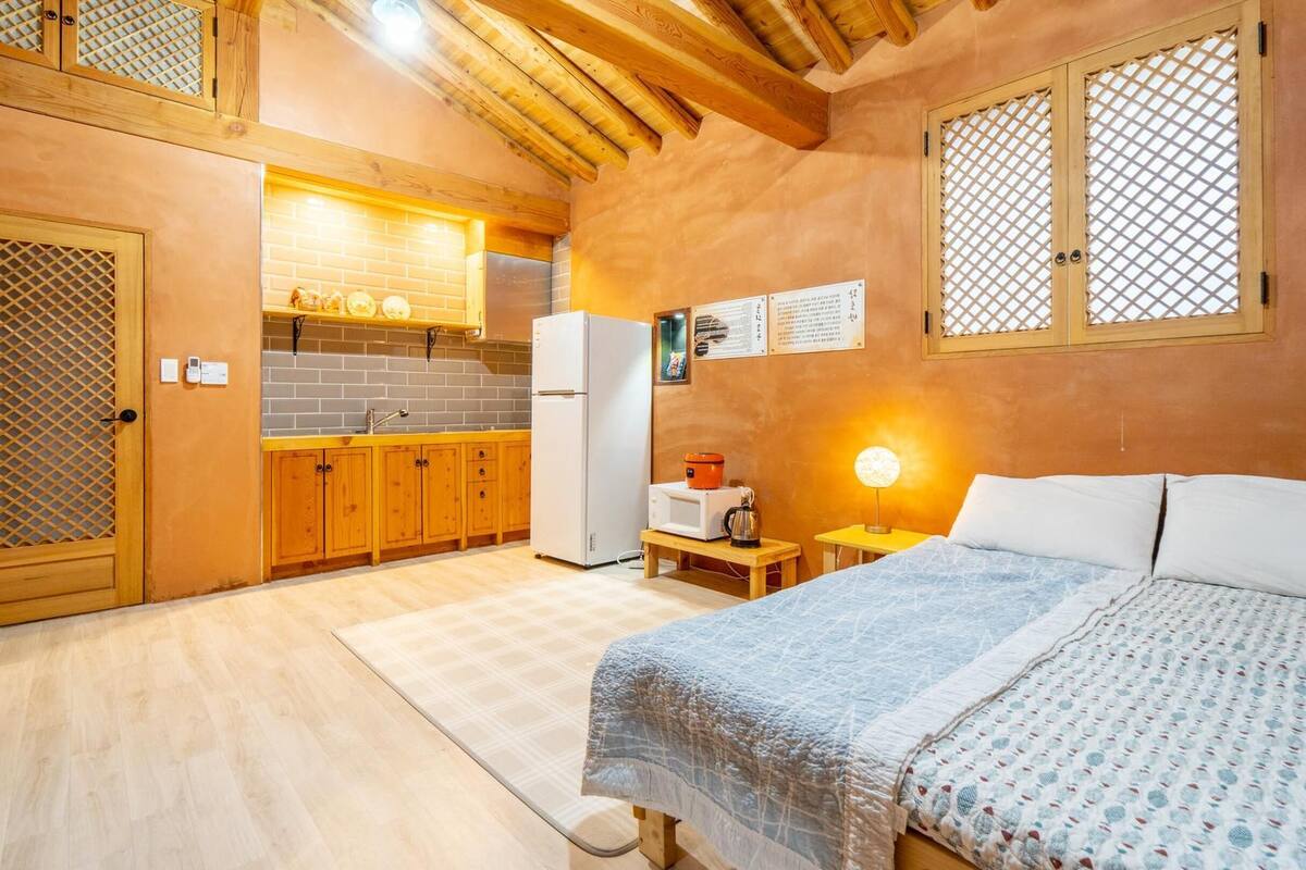 A cozy room with a bed, a small table with a lamp, a kitchen area with a refrigerator, and wooden cabinets.