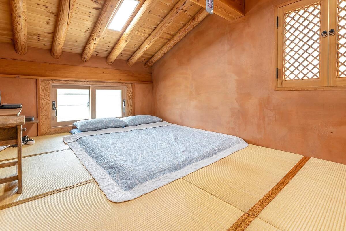 A traditional Japanese-style room with tatami flooring, a low bed, a desk with a computer, and a sliding door with a lattice design.