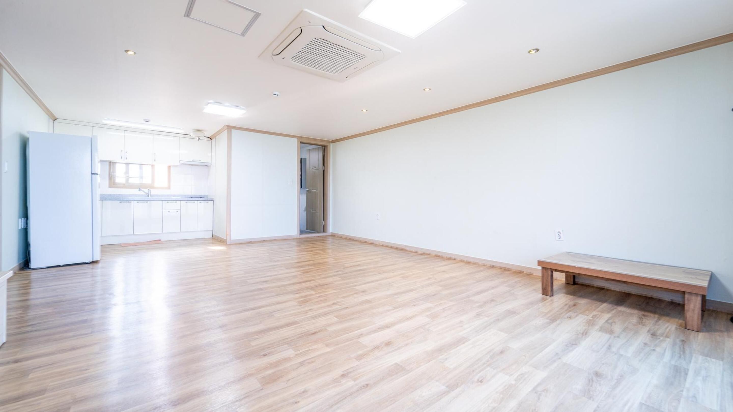 A spacious room with wooden flooring, a bench, and a kitchen area visible through an open door.