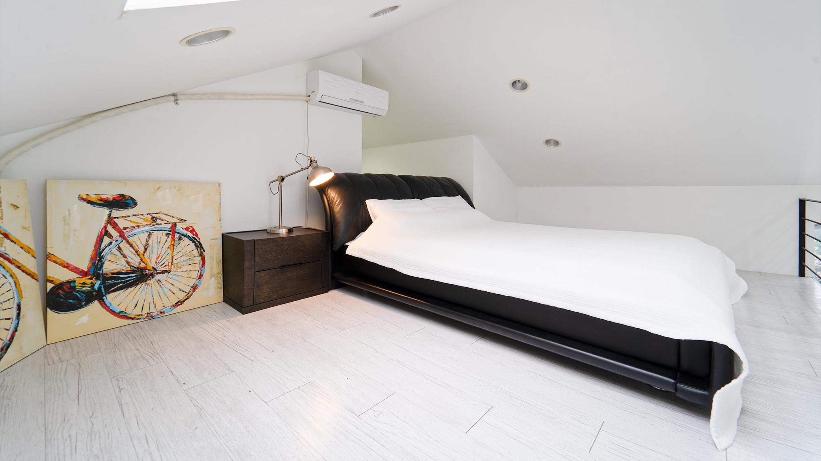 A modern bedroom with a black bed, white bedding, a wooden nightstand, a desk lamp, and a colorful bicycle painting on the wall.