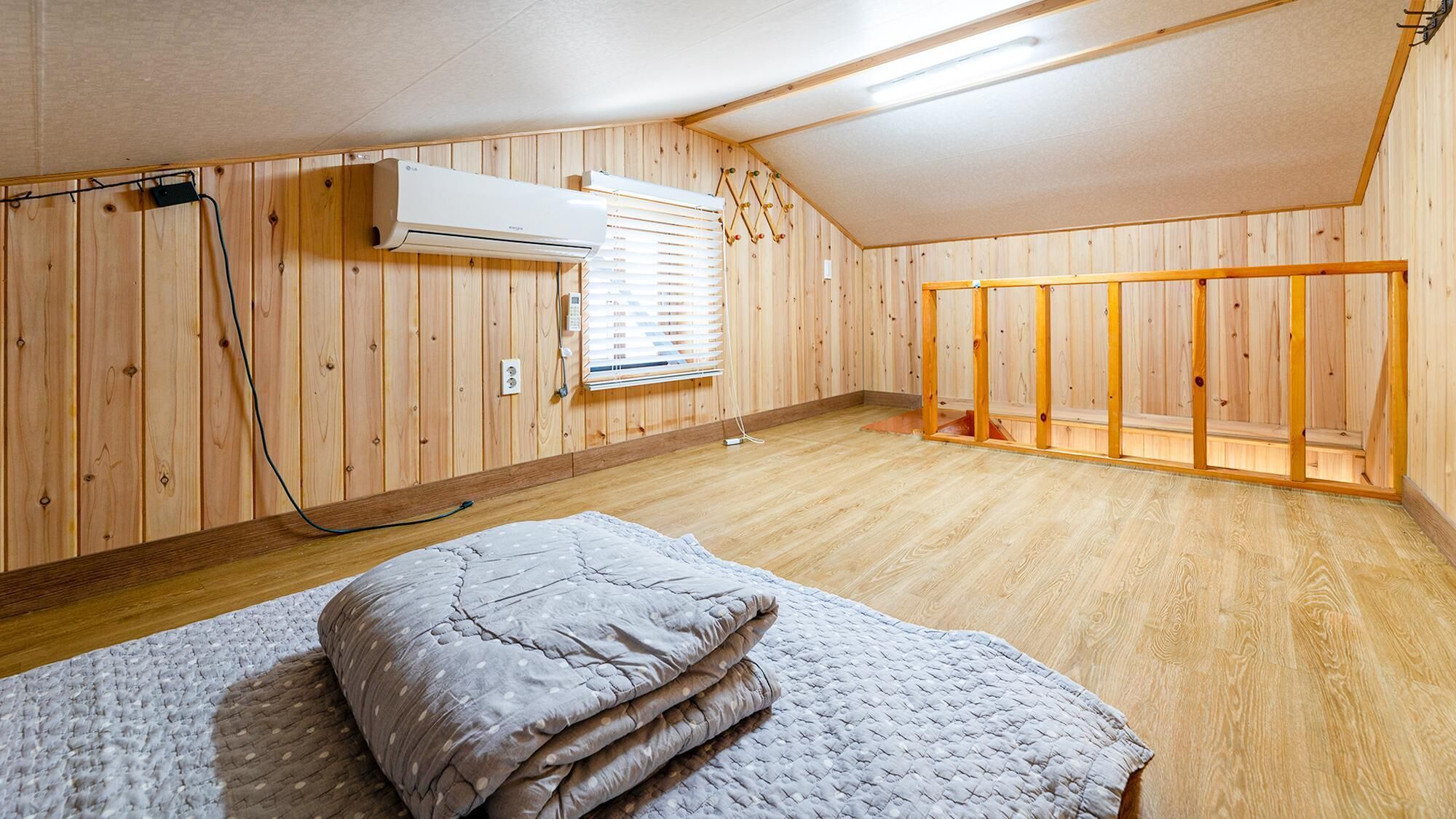 A room with wooden panel walls, a ceiling fan, an air conditioning unit, a bed with a quilted blanket, and a window with blinds.