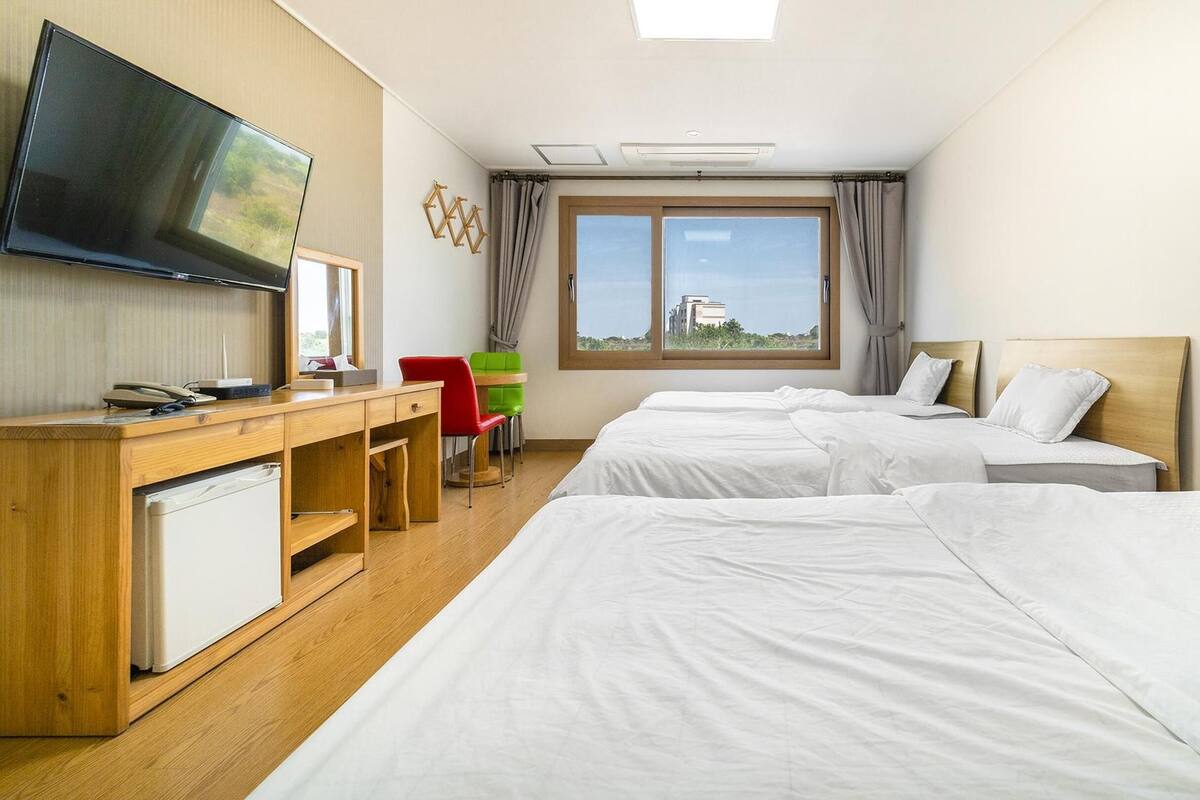 Habitación de hotel con dos camas, un escritorio, televisor y ventana con vistas a edificios.