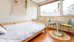 A bedroom with a wooden bed, a round table, two chairs, a macrame wall hanging, and a large window with a view of trees.