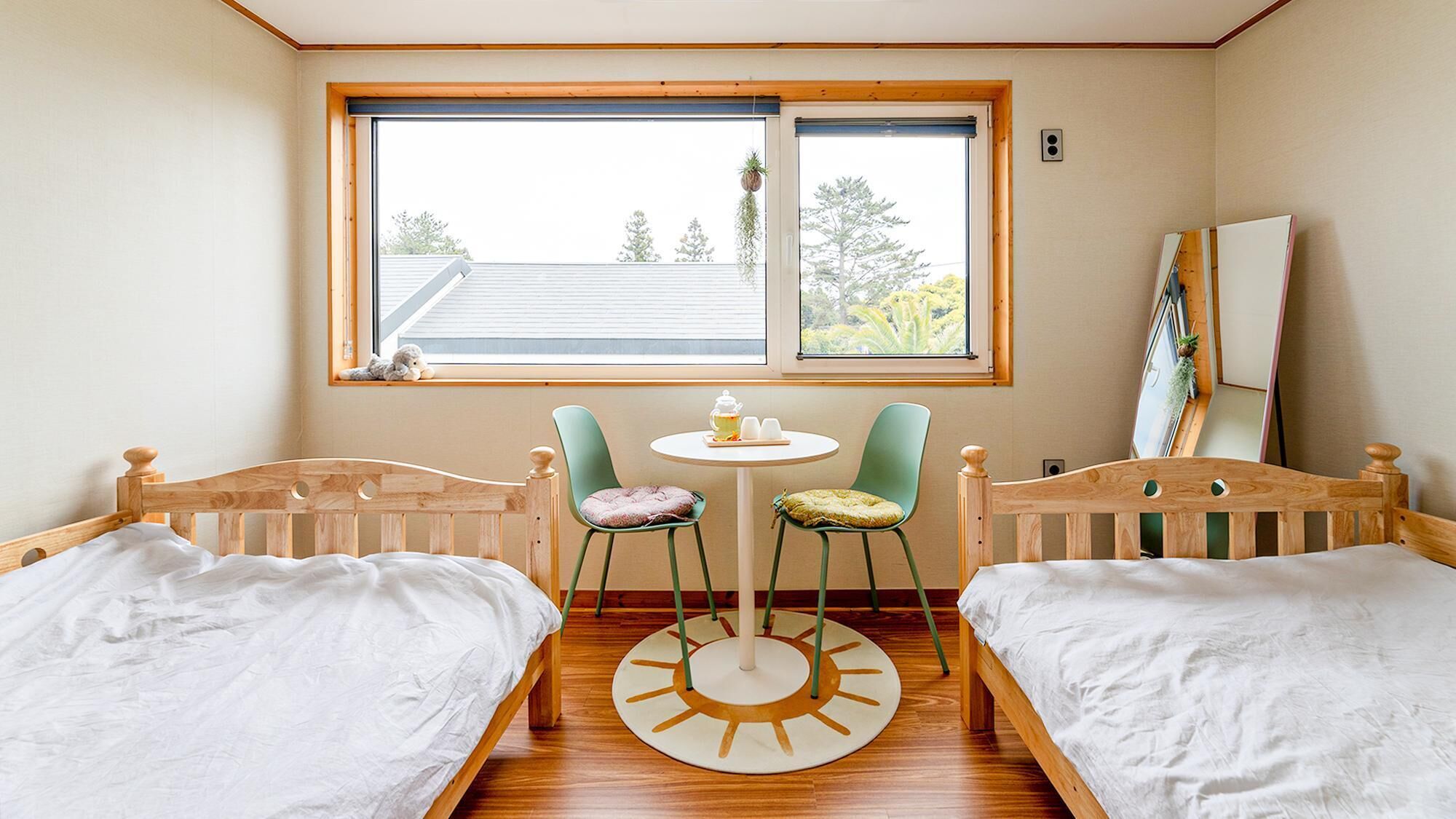 A room with two wooden beds, a small round table with chairs, a window with a view of trees, and a mirror on the wall.