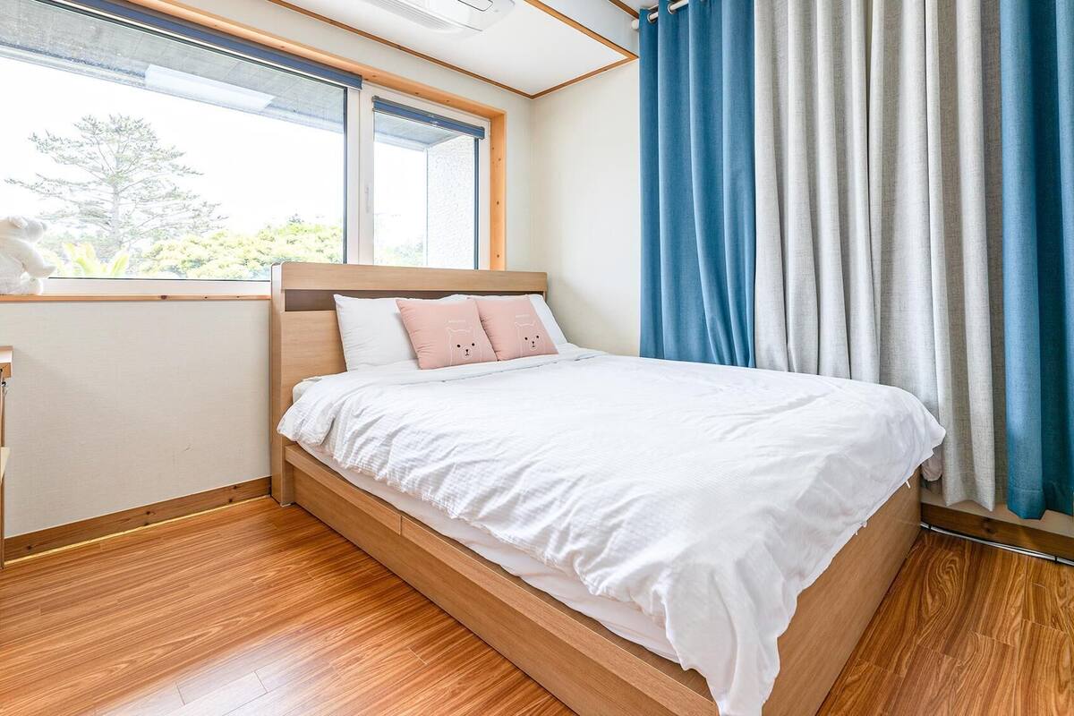 Une chambre à coucher avec un lit en bois, du linge de lit blanc, une fenêtre donnant sur des arbres et des rideaux bleus.
