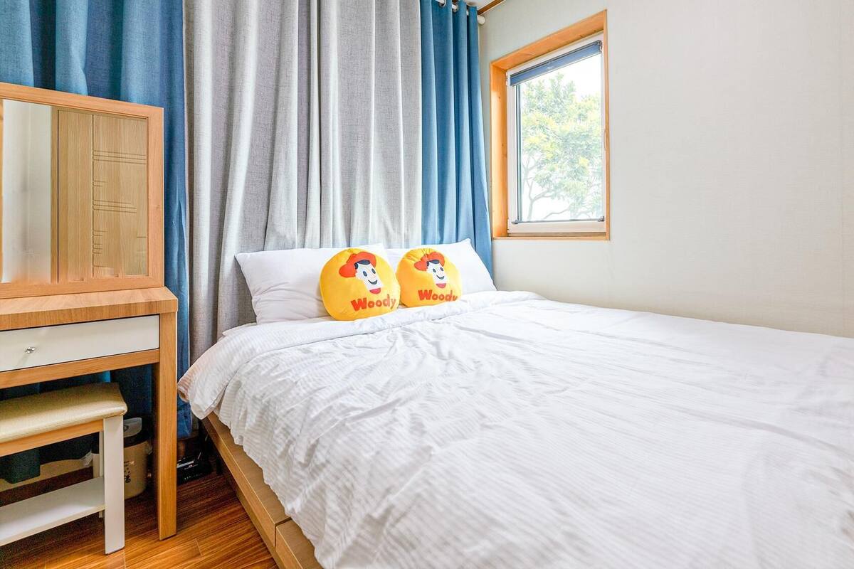 Une chambre avec un lit en bois, du linge de lit blanc, une table de chevet en bois, un miroir et une fenêtre avec des rideaux bleus.