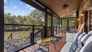 Un espace extérieur abrité doté d’une terrasse en bois, d’une table en verre, d’un canapé avec des coussins à rayures et d’une vue sur une rivière et des arbres.