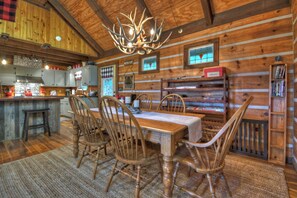 A rustic wooden interior with a dining table set for four, surrounded by wooden chairs. A chandelier hangs from the ceiling, and there are wooden shelves with books and a red box on the wall.