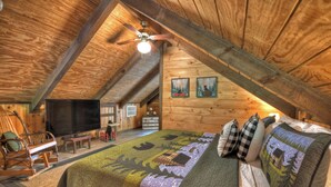 Une chambre confortable dans un chalet en rondins, avec un grand lit, une télévision, un fauteuil à bascule et un ventilateur de plafond.