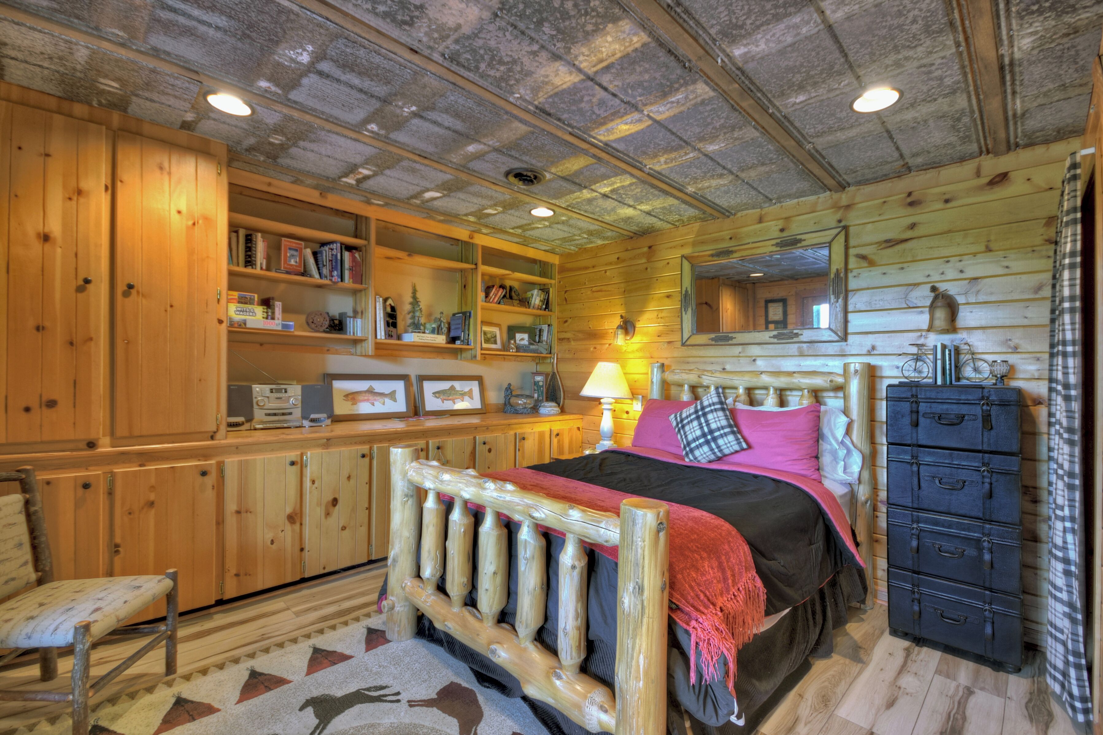 A wooden cabin-style bedroom with a bed, dresser, shelves with books, and a mirror.