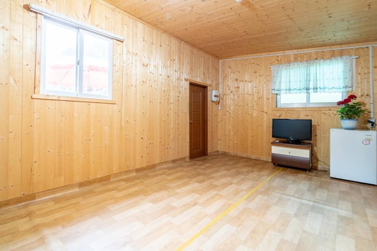 Une pièce aux murs et au sol en bois, avec une fenêtre, une porte, un téléviseur et un réfrigérateur.