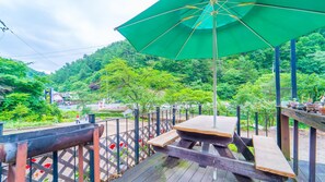 Una terraza de madera con mesas de picnic y un paraguas verde, rodeada de exuberante vegetación y colinas.