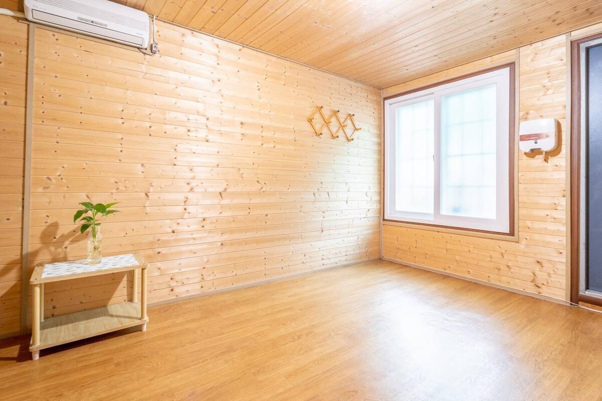 Une pièce aux murs en bois et dotée d’une grande fenêtre. On y trouve une petite table sur laquelle est posée une plante.