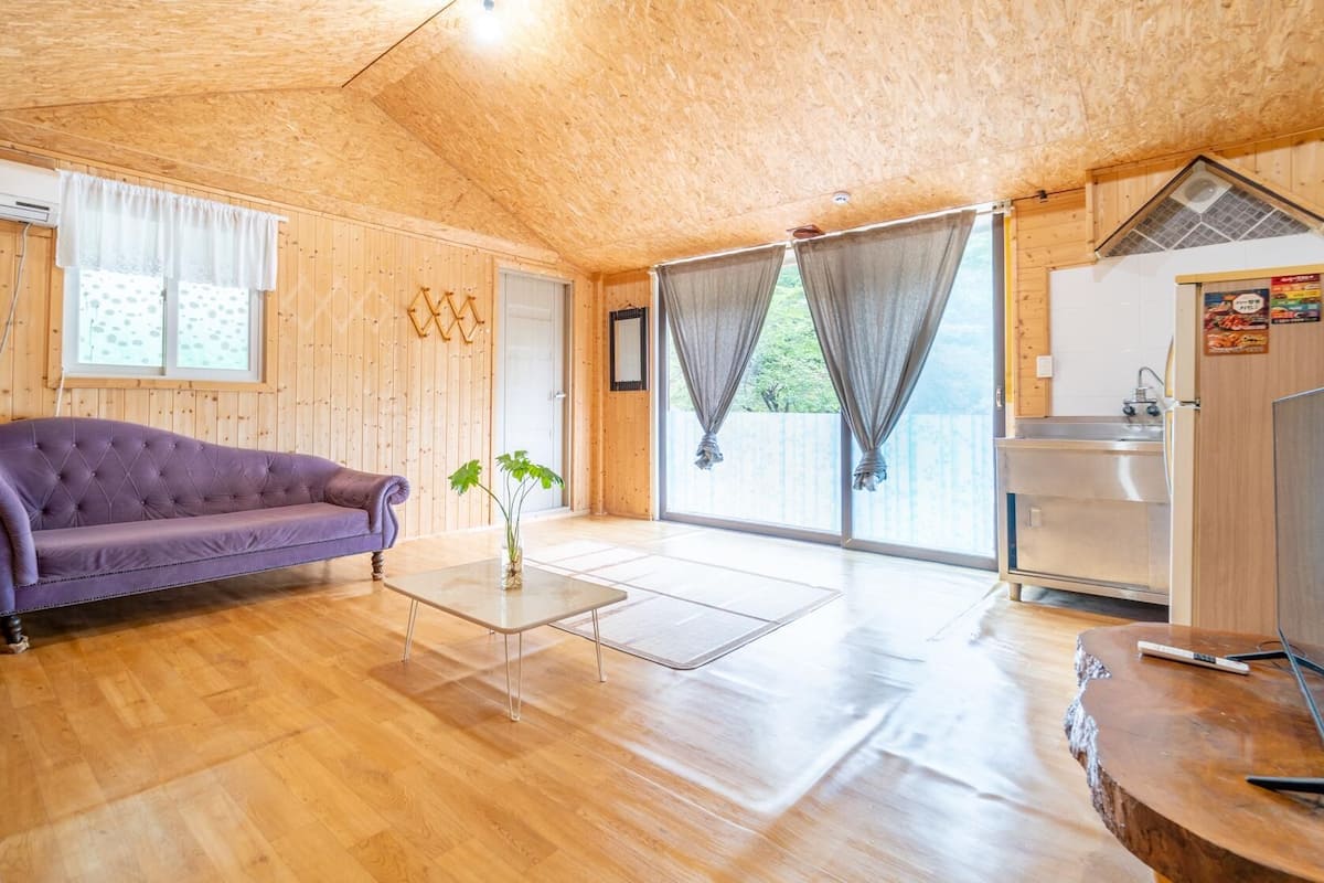 Une cabane en bois chaleureuse, avec un canapé violet, une petite table agrémentée d’une plante et un coin cuisine.
