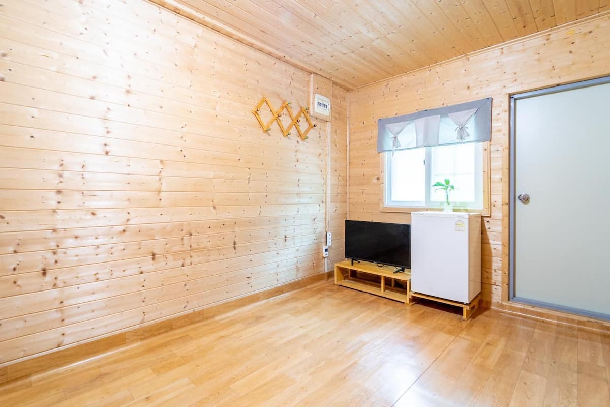 Une pièce de style cabane en bois, avec un plancher en bois, un téléviseur posé sur un meuble en bois, un réfrigérateur blanc et une fenêtre avec des rideaux.