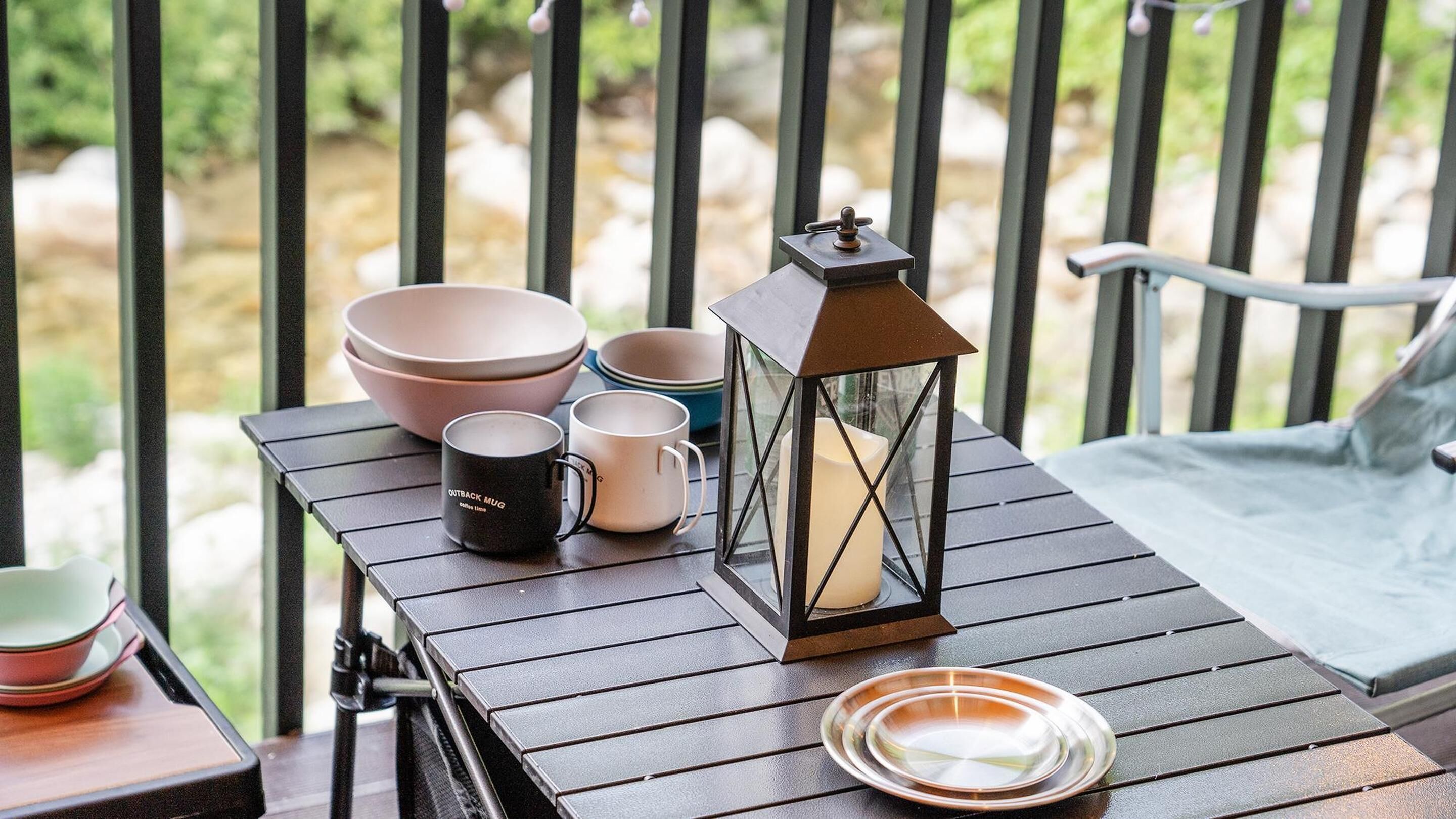 A wooden table with a lantern, bowls, and mugs on a balcony.