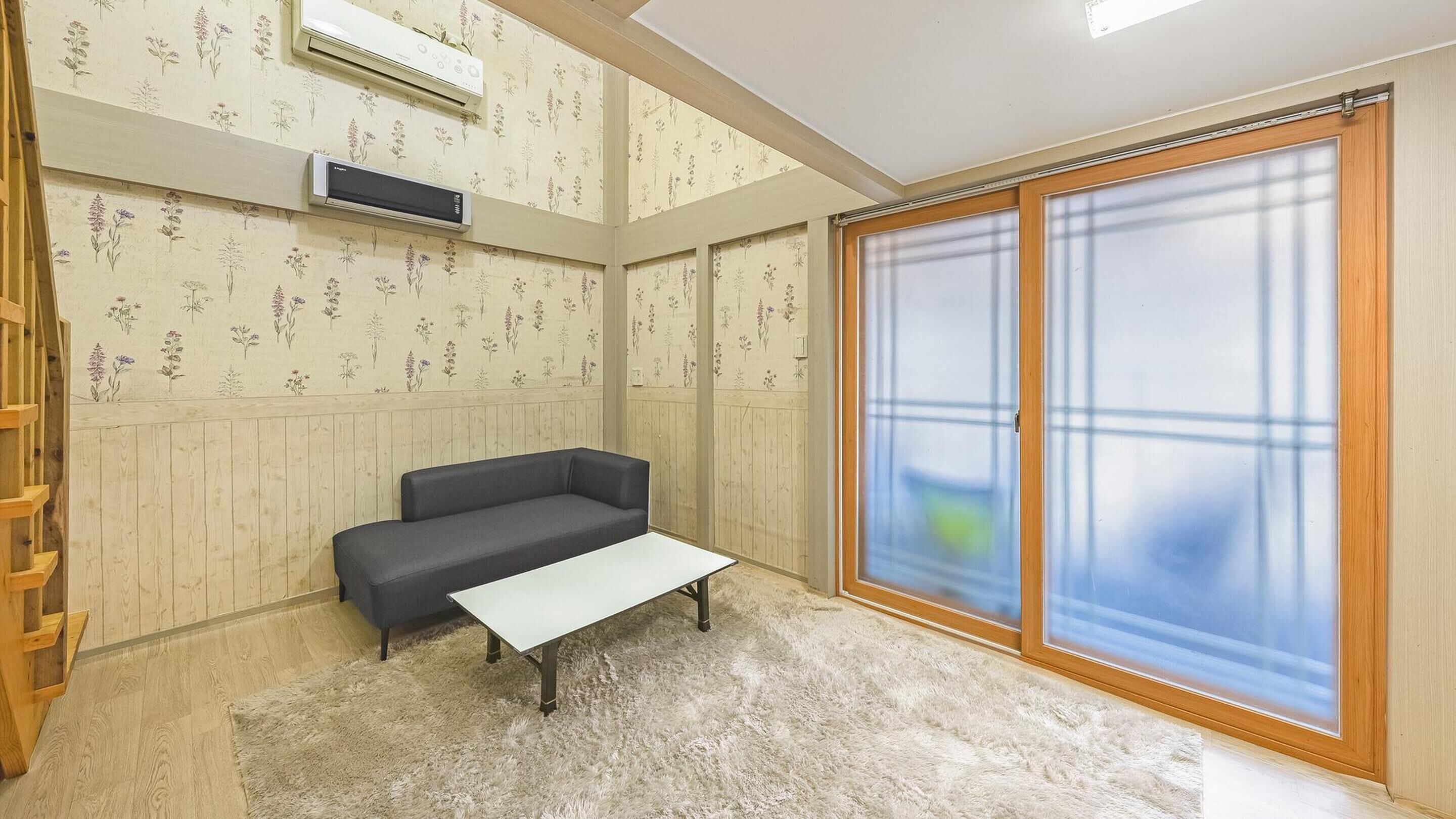 A room with a grey sofa, a white table, wooden flooring, floral wallpaper, and a large sliding glass door with a view.