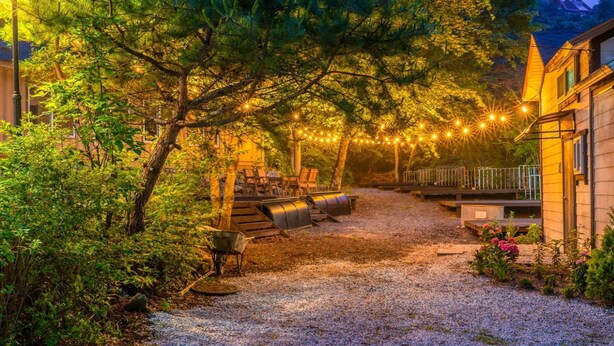 Un camino de grava que lleva a una terraza de madera con mobiliario exterior, rodeado de árboles y luces colgantes.