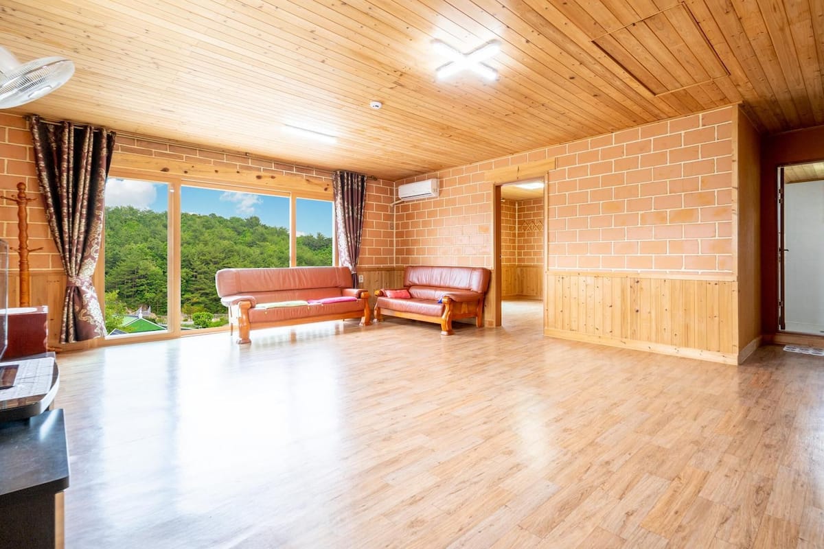 A spacious room with wooden flooring, a leather sofa, and a large window offering a view of greenery.